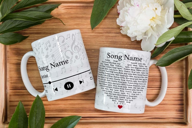 May include: Two white ceramic mugs with musical note designs and the text "Song Name" and "Artist". One mug features a music player interface. The other mug has song lyrics and a red heart. The mugs are on a wooden surface.