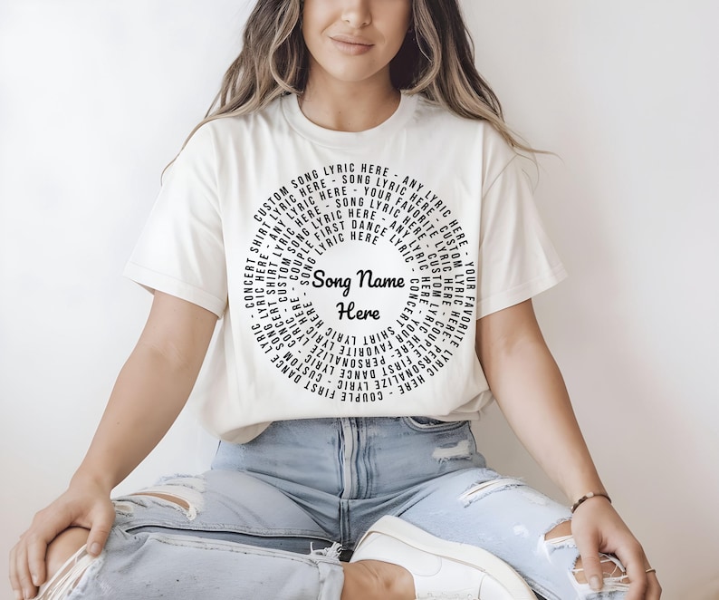 A woman wearing a white t-shirt with a circular design and the text "Song Name Here" printed on it, sitting cross-legged on the floor.