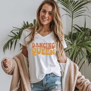 May include: White v-neck t-shirt with the text "DANCING QUEEN" in orange and yellow retro font. The shirt is worn with denim shorts and a tan jacket. The overall theme is casual and retro.