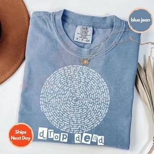 May include: Blue jean colored t-shirt with a circular white text design and the words "drop dead" in white block letters. A gold necklace with a pendant rests on the shirt. The shirt has a Comfort Colors tag.