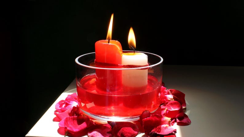 Two candles, one red and one white, tied together with red thread and burning inside a glass bowl filled with red liquid, surrounded by rose petals on a white surface in dramatic lighting. Aesthetic spiritual altar setup featuring candlelight, floral elements, and ritual-themed decor often used in metaphysical or intention-setting photography.
