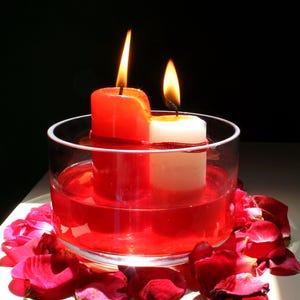 Two candles, one red and one white, tied together with red thread and burning inside a glass bowl filled with red liquid, surrounded by rose petals on a white surface in dramatic lighting. Aesthetic spiritual altar setup featuring candlelight, floral elements, and ritual-themed decor often used in metaphysical or intention-setting photography.