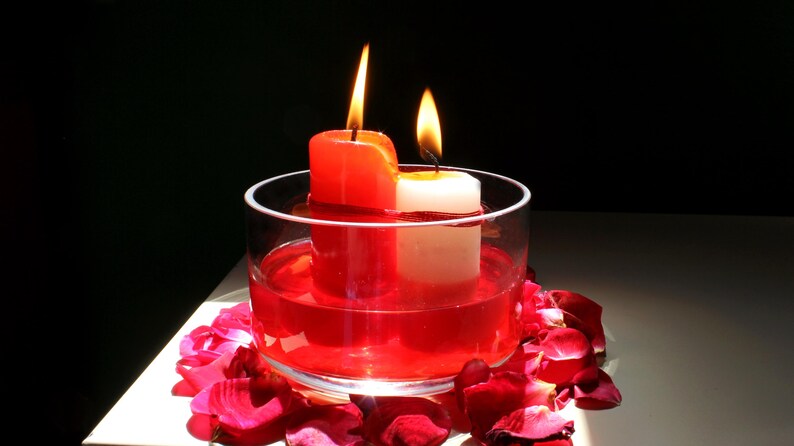 Two candles, one red and one white, tied together with red thread and burning inside a glass bowl filled with red liquid, surrounded by rose petals on a white surface in dramatic lighting. Aesthetic spiritual altar setup featuring candlelight, floral elements, and ritual-themed decor often used in metaphysical or intention-setting photography.