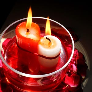 Overhead view of two bound candles, one red and one white, burning together inside a glass bowl filled with red liquid and encircled by rose petals. Warm, dramatic lighting highlights a symbolic ritual altar arrangement used in spiritual imagery, intention-setting photography, candle magic aesthetics, and metaphysical-themed visuals.