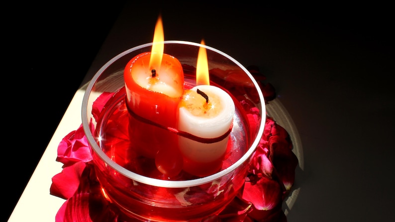 Overhead view of two bound candles, one red and one white, burning together inside a glass bowl filled with red liquid and encircled by rose petals. Warm, dramatic lighting highlights a symbolic ritual altar arrangement used in spiritual imagery, intention-setting photography, candle magic aesthetics, and metaphysical-themed visuals.