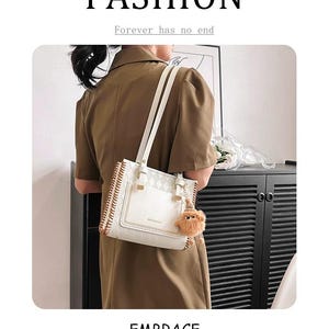 Peut inclure: Un sac à bandoulière blanc avec une texture à motif losanges, doté d'une poche avant, de détails dorés et d'une petite breloque en forme de créature duveteuse marron sur le côté. Le sac possède deux fines anses et des coutures latérales.