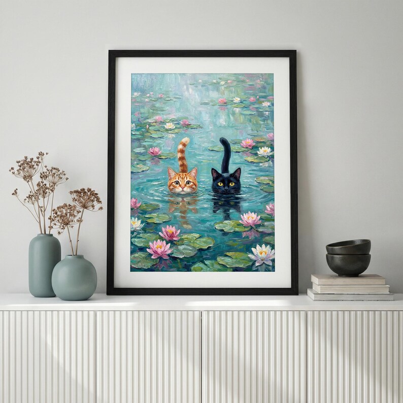 May include: A framed wall art print depicting an orange tabby cat and a black cat swimming in a pond filled with pink and white water lilies and green lily pads. The cats have their heads and tails visible above the water surface.