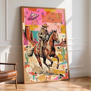 May include: A framed wall art print featuring a colorful illustration of a cowboy riding a horse through a desert landscape with cacti and rock formations. Above the rider, a flying saucer hovers in a pink sky filled with stars and a crescent moon.