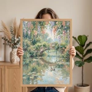 May include: A framed landscape painting depicts a serene pond scene with a stone bridge, lush greenery, and colorful hanging flowers. A duck swims in the water, reflecting the sky and surrounding foliage. The frame is made of light-colored wood.