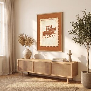 May include: A rectangular wall art print featuring a terracotta-colored background with a central cream-colored postage stamp design. The stamp depicts three figures in wide-brimmed hats riding horses in a line, rendered in a dark brown outline.