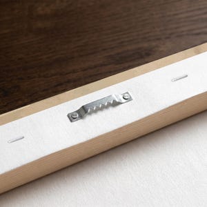 May include: A rectangular wooden frame with a white canvas backing. A silver-colored metal sawtooth hanger is securely attached to the center of the wooden frame with two small nails. The canvas is pulled taut and fastened with staples.