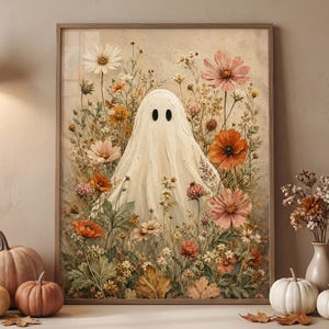 May include: A framed wall art print featuring a central white ghost figure with two black oval eyes, surrounded by a dense arrangement of colorful wildflowers including orange, pink, and white blossoms, set against a muted beige background.