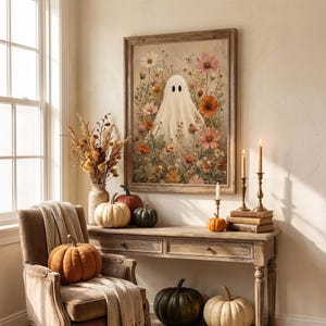 May include: A framed wall art print featuring a white ghost surrounded by colorful wildflowers, displayed above a wooden console table with pumpkins, a vase of dried botanicals, and three lit taper candles in brass holders.