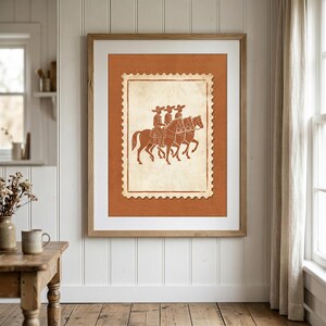 May include: A framed wall art print featuring a postage stamp design with a terracotta-colored background. The central illustration depicts three figures wearing wide-brimmed hats riding horses in profile, rendered in a matching terracotta line art style.