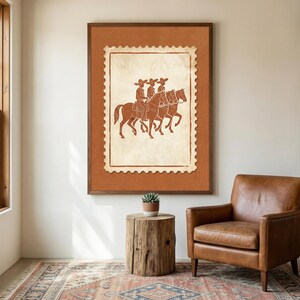 May include: A framed wall art print featuring a postage stamp design with a terracotta-colored background. The central illustration depicts three figures wearing wide-brimmed hats riding horses in profile, rendered in a dark brown line art style.