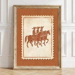 May include: A framed wall art piece featuring a stamp-shaped illustration of three riders on horseback wearing wide-brimmed hats. The artwork uses a terracotta and cream color palette, set against a textured orange background within a wooden frame.