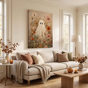 May include: A rectangular wall art print featuring a white ghost figure centered among a variety of colorful wildflowers in shades of orange, pink, and white. The artwork is displayed on a light-colored wall above a beige sofa with throw pillows.