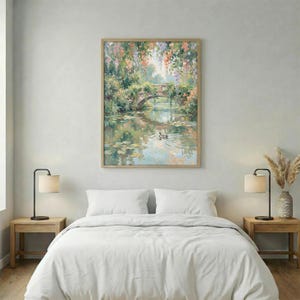 May include: A framed landscape painting depicts a stone bridge over a calm body of water, with ducks swimming. Lush greenery and colorful flowers frame the bridge. The artwork is in a light wooden frame and hangs above a bed with white bedding.