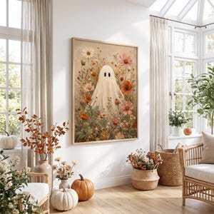 May include: A framed wall art print featuring a central white ghost figure surrounded by a dense arrangement of colorful wildflowers in shades of orange, pink, and cream, set against a soft, neutral-toned background with visible brushstrokes.