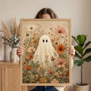 May include: A framed rectangular wall art print featuring a central white ghost figure with two black oval eyes, surrounded by a dense arrangement of wildflowers in shades of orange, pink, cream, and green, set against a textured beige background.