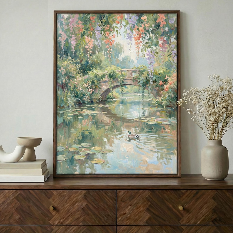 May include: A framed oil painting depicts a serene garden scene. A stone bridge arches over a calm pond with lily pads. Colorful flowers and foliage frame the bridge, and a duck swims in the water. The painting is in a dark wooden frame.