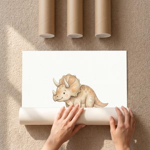 Puede incluir: Ilustraci&oacute;n en acuarela de un simp&aacute;tico dinosaurio Triceratops en tonos beige y marr&oacute;n. La obra de arte est&aacute; sobre papel blanco, parcialmente enrollado, con tres tubos de cart&oacute;n marr&oacute;n encima.
