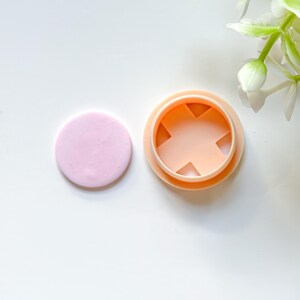 Può includere: Un pezzo di argilla rotondo rosa chiaro accanto a un tagliabiscotti color pesca con un design a forma di X all'interno. Gli oggetti sono su una superficie bianca, suggerendo un tema artigianale o di cottura.