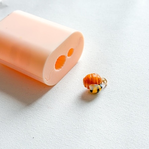 Può includere: Un piccolo ciondolo a forma di ape arancione e nero con un anellino dorato, posizionato accanto a uno stampo in silicone color pesca pallido caratterizzato da una cavità cilindrica e un'apertura circolare più piccola sulla superficie piatta.