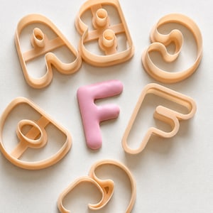 Può includere: Una collezione di tagliabiscotti in plastica color pesca chiaro a forma di lettere A, B, C, D e F, disposti attorno a una singola lettera F solida, liscia e rosa chiaro, realizzata in pasta di zucchero o argilla, posizionata al centro.