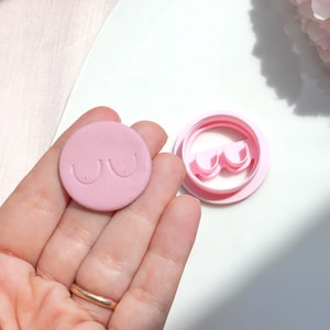 Peut inclure: Un emporte-pièce rond rose avec un motif de sein stylisé. Le découpoir est tenu dans une main, à côté d'un découpoir rose assorti qui forme la forme. Le design comprend deux lignes courbes et deux petits points.