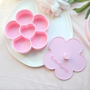 May include: A pink flower-shaped container with a heart-shaped center and six compartments. The lid, also flower-shaped, has a small heart-shaped handle and the text "CutterByMila". The container sits on a white plate.