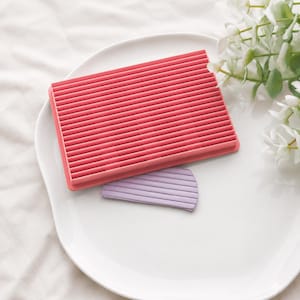 Può includere: Una piastra per texture rettangolare in plastica rosa caratterizzata da una serie di creste orizzontali parallele sulla superficie, accompagnata da un piccolo pezzo curvo di argilla viola chiaro con un motivo a coste impresso sopra.
