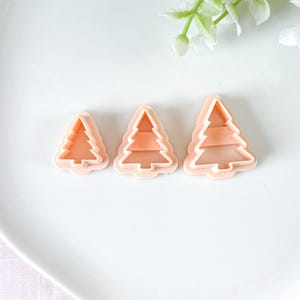 Op de afbeelding: Drie lichtperzik-kleurige kerstboomvormige koekjessnijders. Elke snijder heeft een witte omtrek en een gedetailleerd boomontwerp. De snijders zijn in een rij gerangschikt, variërend in grootte van klein tot groot.