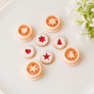 May include: A collection of round cookie cutters in light peach plastic, each featuring a central shape including a heart, snowflake, and tree, alongside three baked round biscuits with red jam centres and icing sugar dusting.
