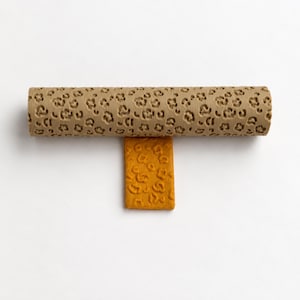 Può includere: Un mattarello cilindrico beige con un motivo leopardato in rilievo ripetuto, posizionato orizzontalmente sopra un pezzo rettangolare di argilla giallo senape che mostra la stessa texture leopardata impressa sulla sua superficie.