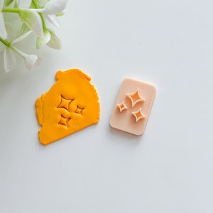 May include: A light pink rectangular stamp featuring three starburst designs. Beside it is a piece of orange clay with the same starburst impressions. The items are on a white surface.