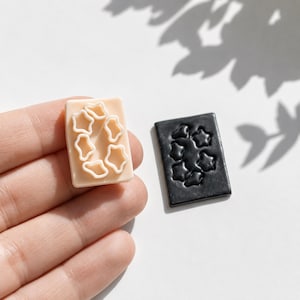 May include: A rectangular peach-coloured plastic cookie cutter featuring a raised star pattern and a matching rectangular black clay slab with an impressed star design, showcasing the tool and its resulting embossed shape for crafting projects.