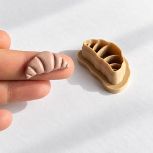 Könnte beinhalten: Ein kleines, hellbraunes, halbmondförmiges Objekt im Croissant-Stil mit strukturierter Oberfläche, platziert neben einem passenden beigen Kunststoff-Ausstecher, der ein hohles, geschichtetes Design zum Formen von Teig in Croissant-Form aufweist.
