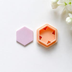 May include: A light purple hexagonal flat piece and a peach-coloured hexagonal cutter with an internal cross-shaped cutout. The cutter features a raised rim and a hollow centre, while the flat piece is solid and smooth, both designed for crafting.
