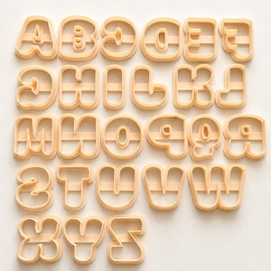 Pode incluir: Uma coleção de vinte e seis cortadores de biscoitos de plástico na cor pêssego claro, moldados como as letras do alfabeto de A a Z. Cada letra apresenta um contorno espesso e arredondado e uma barra horizontal central para suporte estrutural.