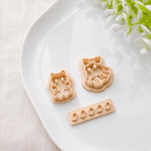 May include: Two light beige plastic cookie cutters shaped like cartoon characters with bows, accompanied by a rectangular strip containing five small, uniform ghost-shaped cutters, all featuring raised edges for precise dough impressions.