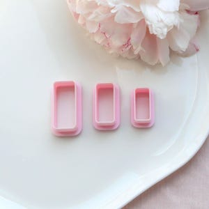 Può includere: Tre formine per biscotti rettangolari rosa di diverse dimensioni sono disposte su un piatto bianco. Sullo sfondo una grande peonia rosa chiaro. Le formine sono in plastica e hanno una finitura liscia.