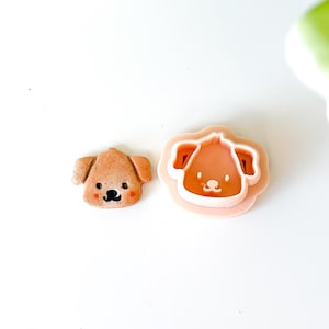 Peut inclure: Un biscuit en forme de chien cuit, de couleur brun clair, à côté d'un emporte-pièce en forme de chien. Le biscuit a un nez et des yeux noirs, avec des joues roses. L'emporte-pièce est rose clair avec un intérieur blanc.