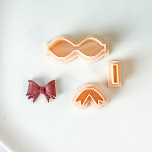 May include: A selection of bow-shaped cookie cutters and a finished bow. The cutters are light peach-coloured plastic, and the bow is a dark red with glitter. The cutters are in various shapes and sizes.