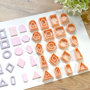 May include: An assortment of geometric polymer clay cutters in various shapes, including squares, circles, and triangles. The cutters are peach-colored, and the clay pieces are shades of pink and lavender. The items are arranged on a white surface.