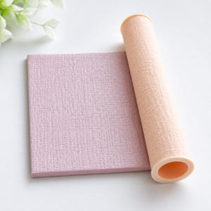May include: A rectangular mauve sheet with a textured surface placed next to a cylindrical peach-colored tube featuring a matching fine-grained texture. Both items exhibit a consistent matte finish and are positioned side by side on a plain surface.