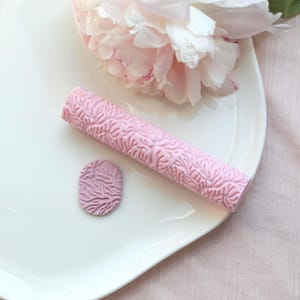 Op de afbeelding: Een roze, getextureerde deegroller en een klein ovaal stuk met hetzelfde patroon liggen op een wit bord. Op de achtergrond een grote, lichtroze pioenroos. De deegroller heeft een herhalend, organisch ontwerp.