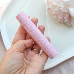 Peut inclure: Un objet cylindrique rose clair et texturé est tenu dans une main. L'objet repose sur une assiette blanche, avec une fleur rose en arrière-plan. Une bague dorée est visible sur la main.