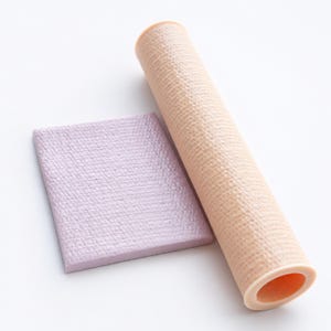 Op de afbeelding: Een lichtbeige cilindrische buis met een getextureerd oppervlak naast een plat, rechthoekig lavendelkleurig vel met een bijpassend reliëfpatroon. Beide items vertonen een consistente, fijnkorrelige, kruislings gearceerde textuur.