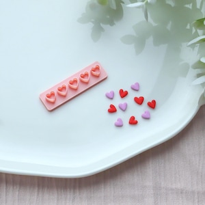 Könnte beinhalten: Eine rosa rechteckige Form mit sechs herzförmigen Aussparungen, neben verstreuten roten und lila herzförmigen Streuseln. Die Artikel sind auf einem weißen Teller mit gewelltem Rand angeordnet, was auf ein Back- oder Bastelthema hindeutet.